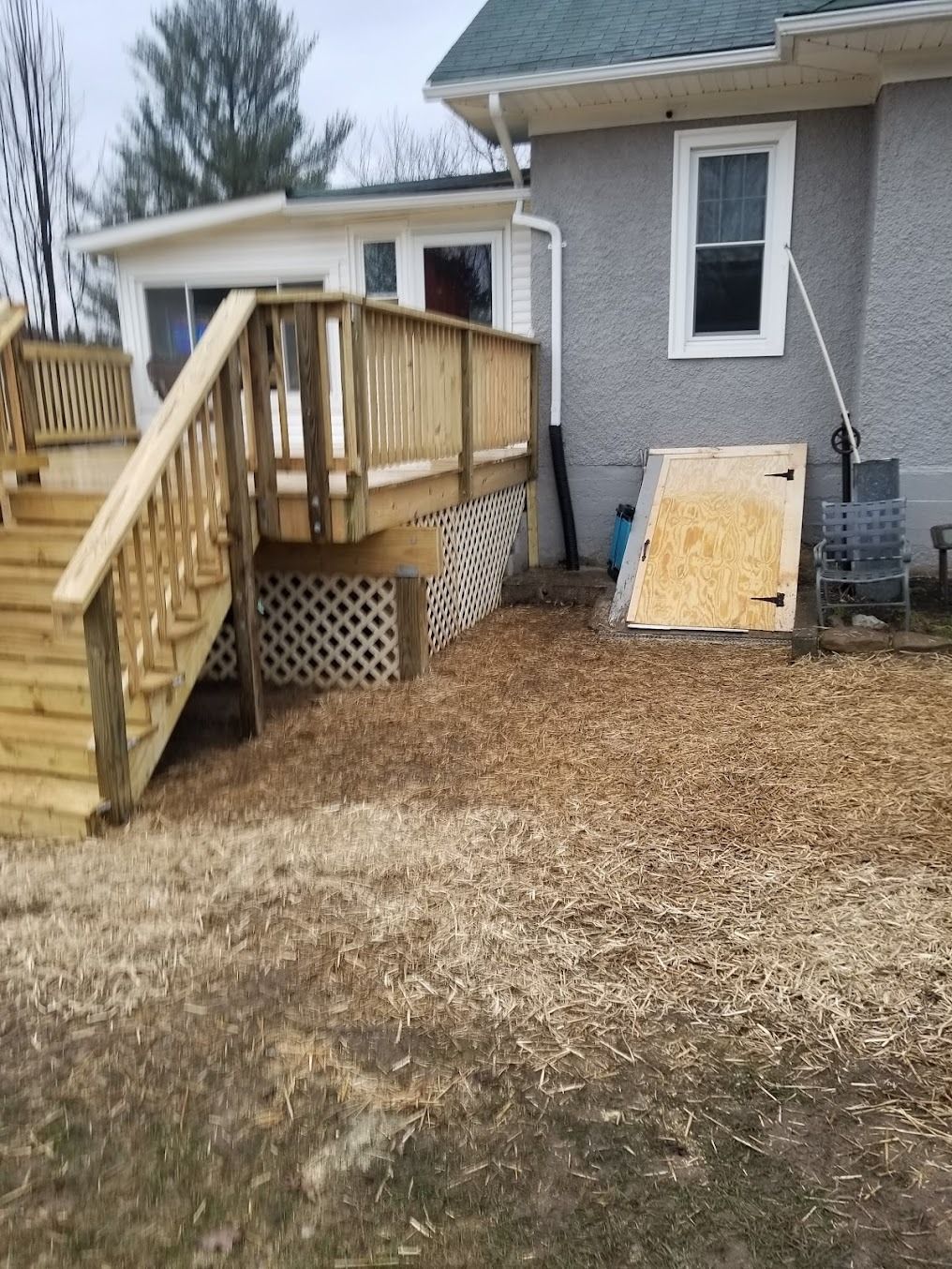 Wooden wheelchair ramp to a gray house, with plywood leaning against the wall and wood chips on the ground.