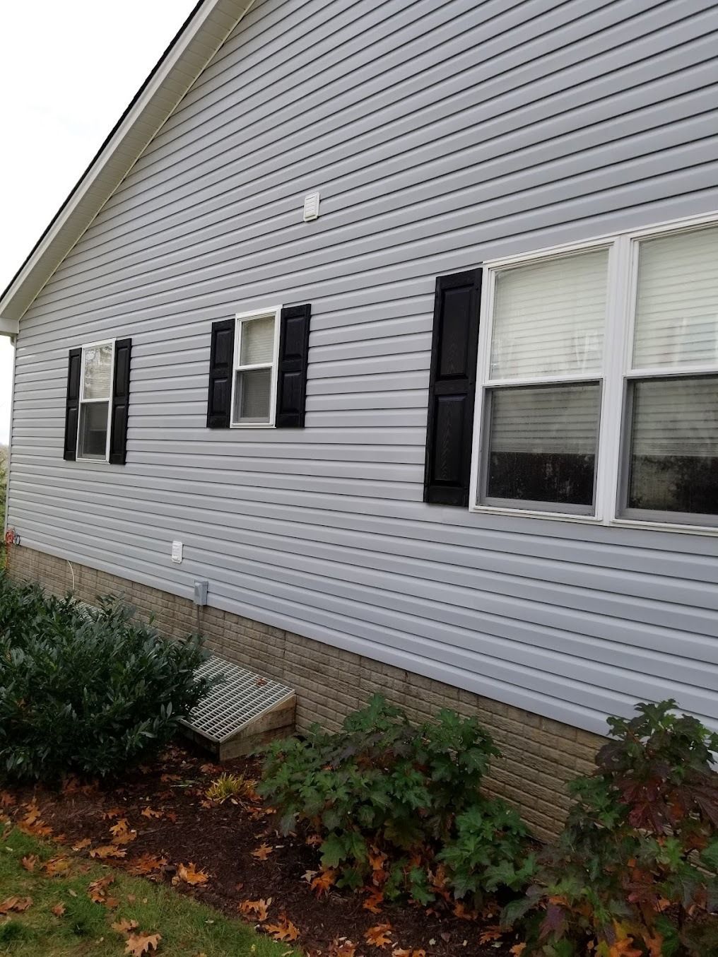 Side of a gray house with black shutters and white-trimmed windows, beside shrubs and wet ground.