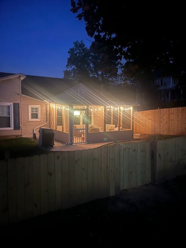 Backyard patio at dusk with warm string lights on a covered porch and wooden fence in front.