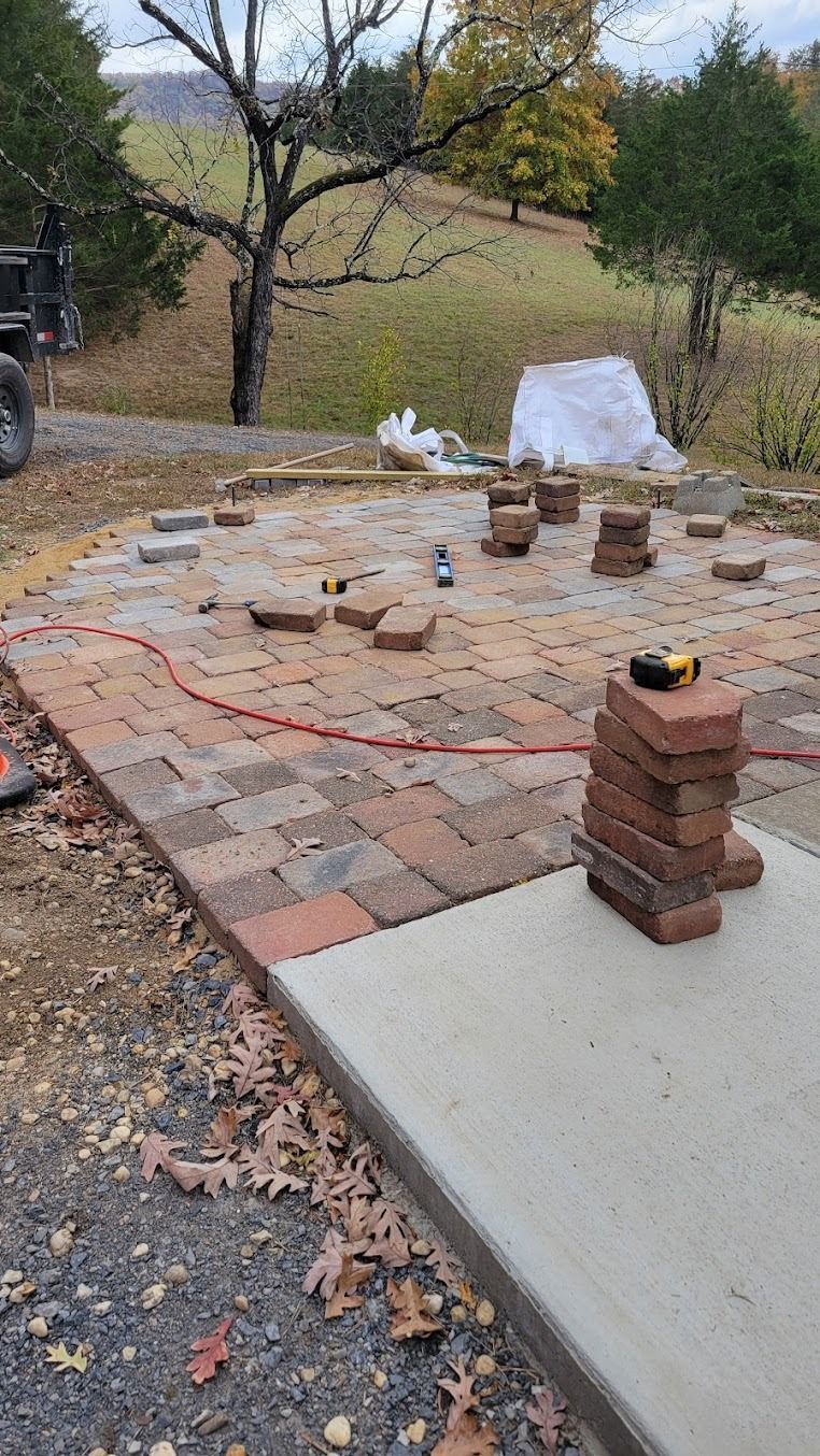 Partially built brick patio with scattered pavers and a small brick stack beside a concrete slab outdoors