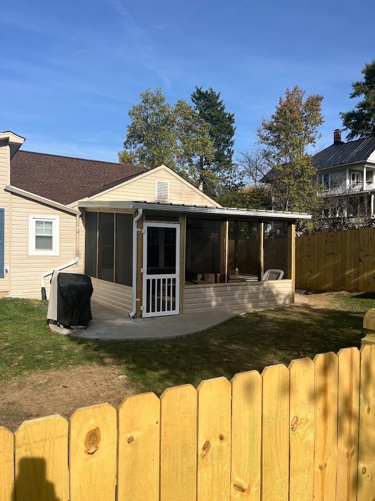 Backyard patio with a small covered porch, beige house, and yellow wooden fence under a blue sky