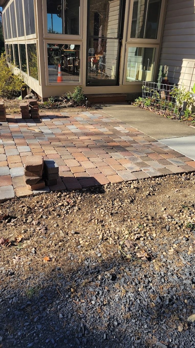 Brick patio and gravel path outside a house with large windows and shrubs.
