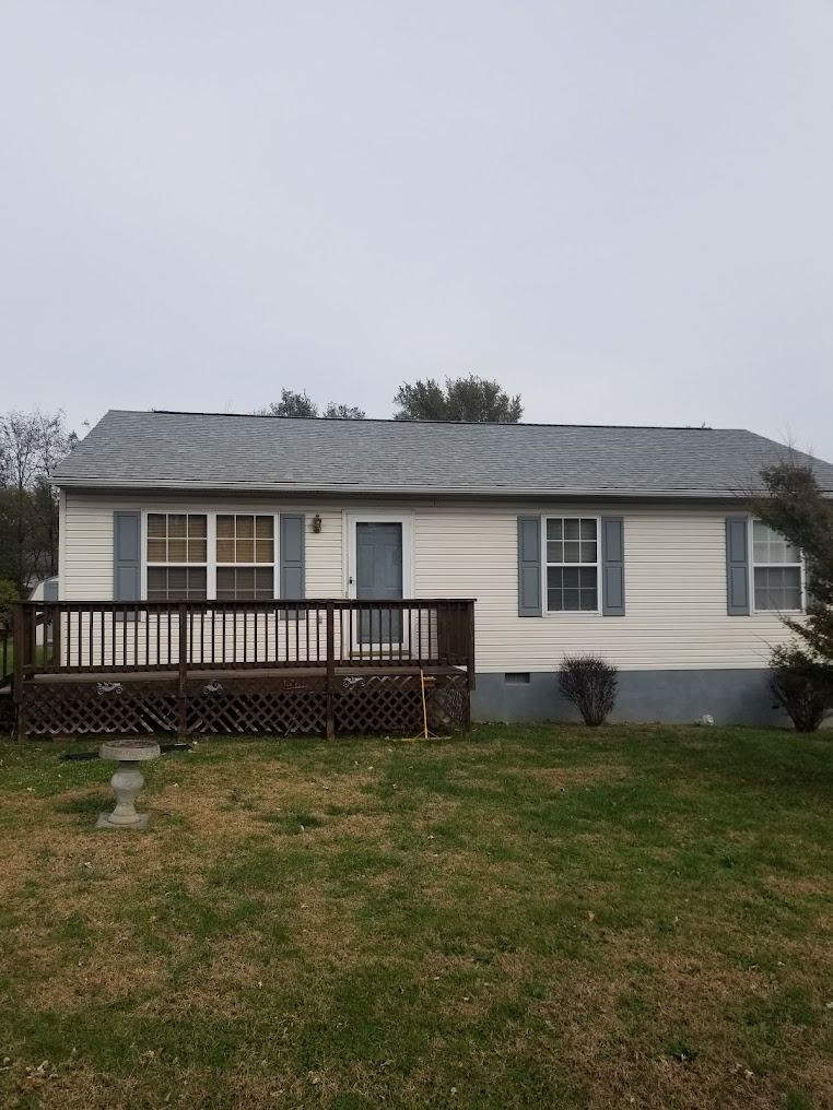 Single-story beige house with gray roof, front deck, and patchy lawn under an overcast sky