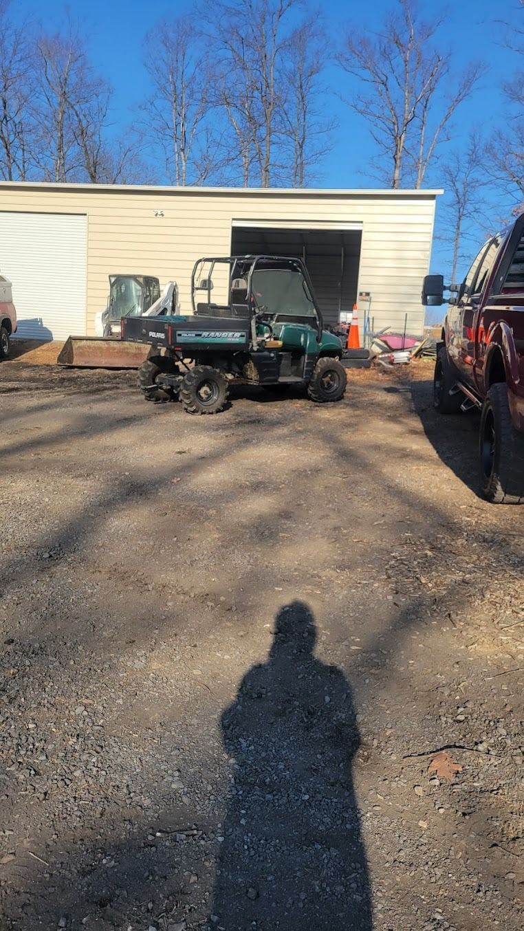 Utility vehicle parked in front of a beige building, with a truck at right and long shadows on gravel.