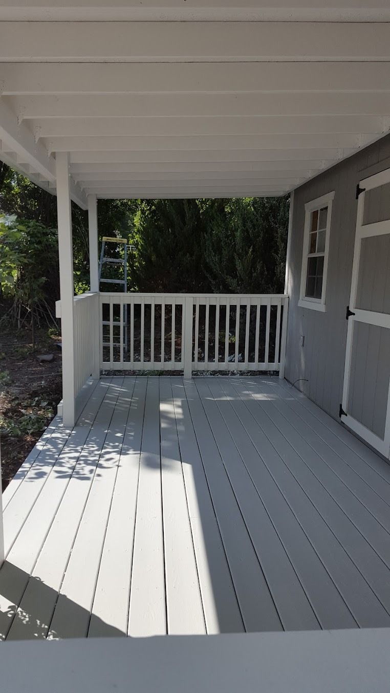 Covered porch with white railing, gray siding, and a sunlit painted floor.