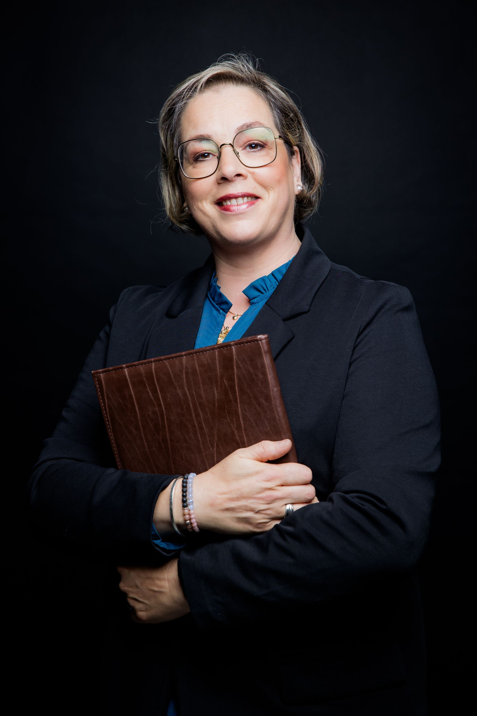 A smiling person with glasses, wearing a teal long-sleeved blouse, poses against a solid yellow background.