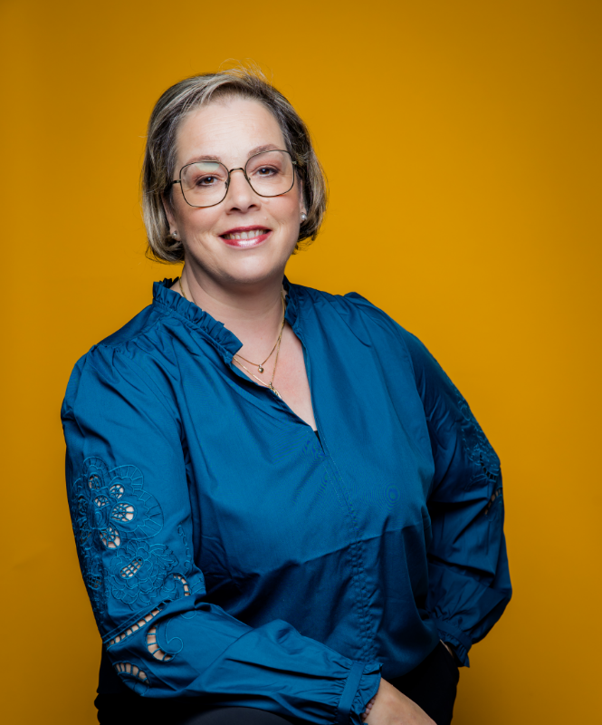 A smiling person with glasses, wearing a teal long-sleeved blouse, poses against a solid yellow background.