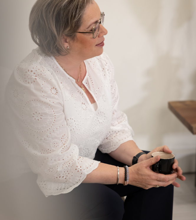 A person wearing a white eyelet blouse and glasses sits in profile, holding a dark mug while looking off to the side.