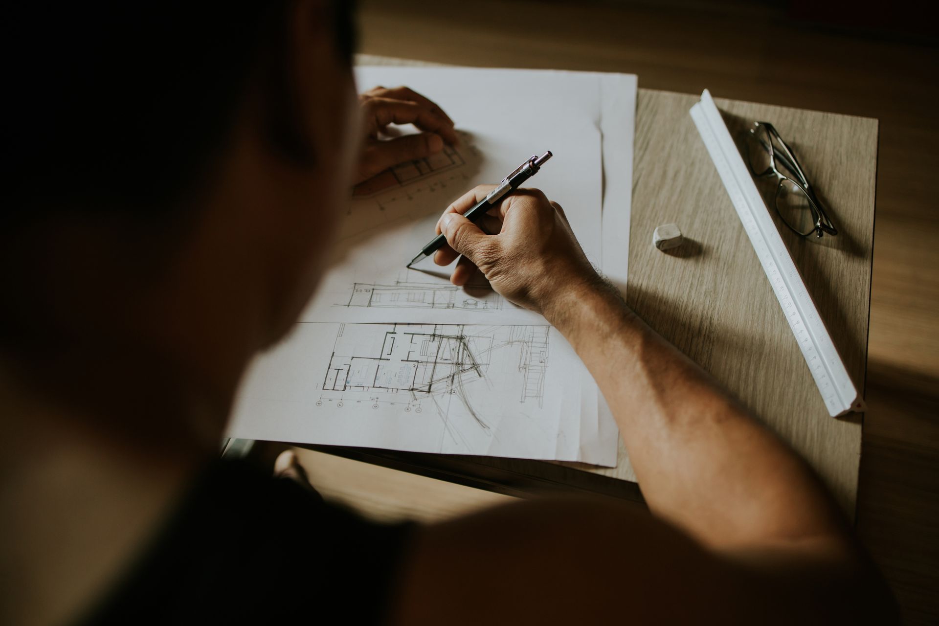 Person sketching a floor plan on paper at a desk, with tools and a ruler nearby.