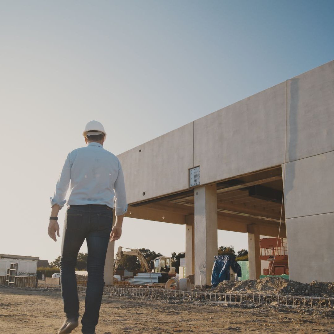 Person in a hard hat walks toward a large concrete building under construction.