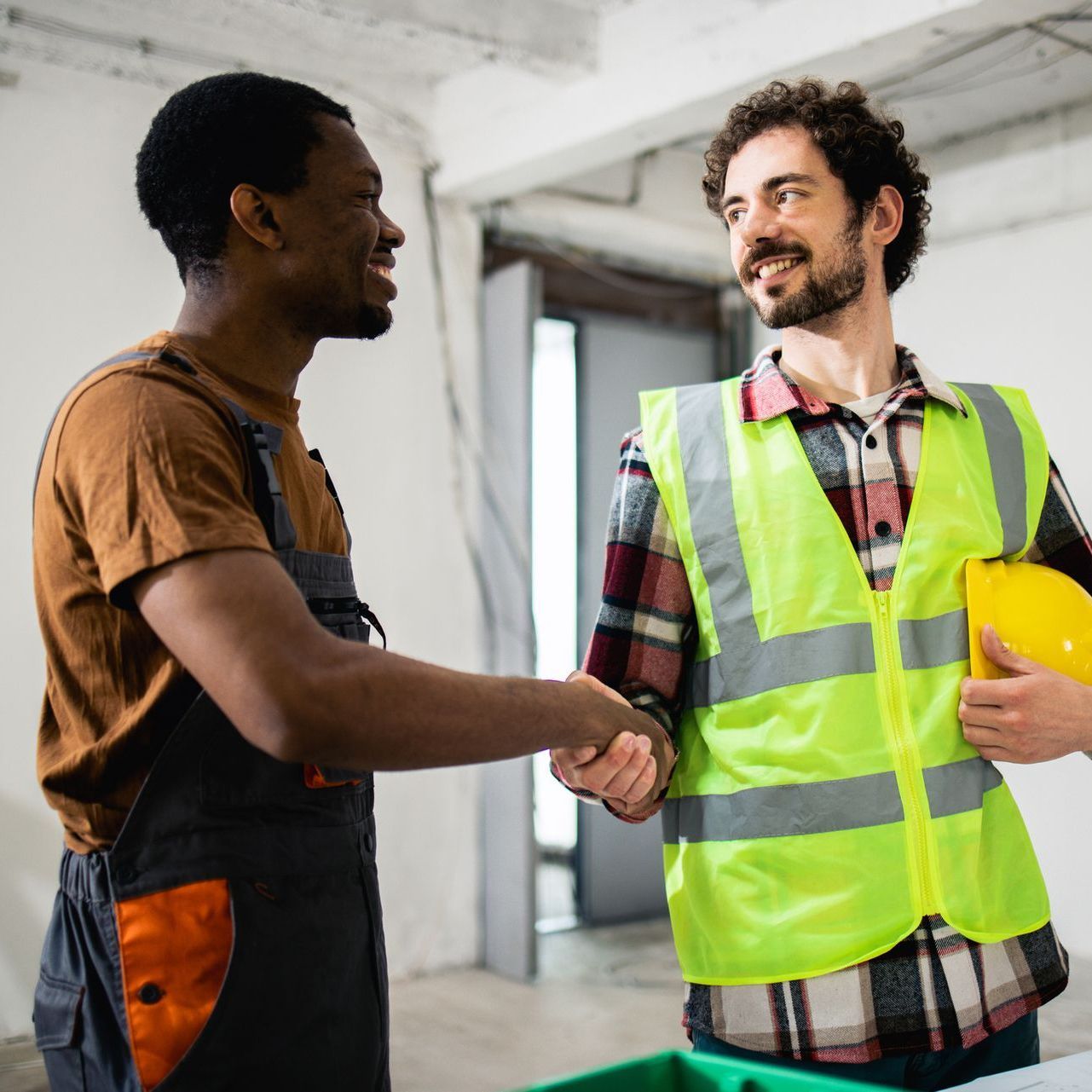 Two workers shake hands, one in a neon safety vest and hard hat, in a warehouse.