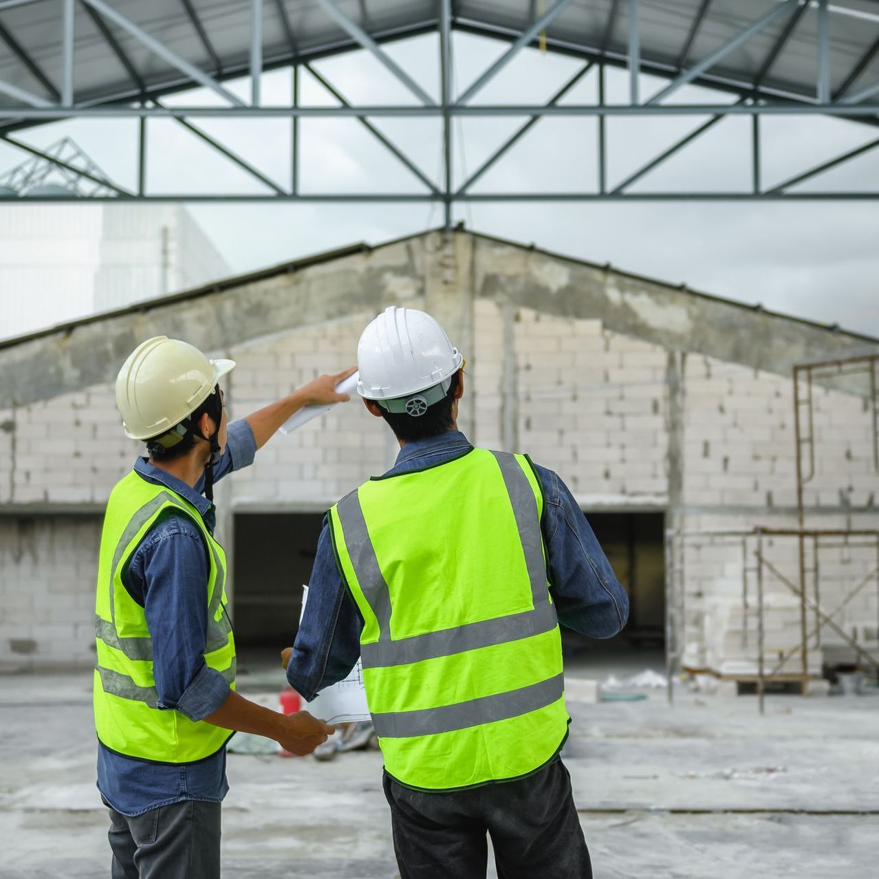 Two construction workers in safety vests and hard hats inspect a building site