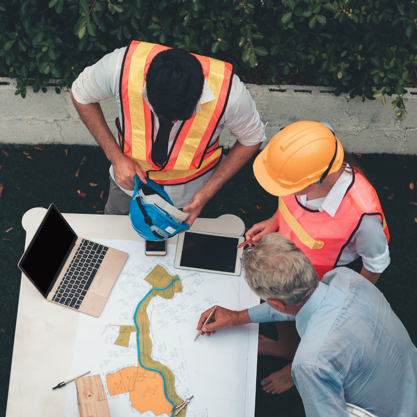 Three people in safety vests review a map at a table with a laptop and tablet outdoors.