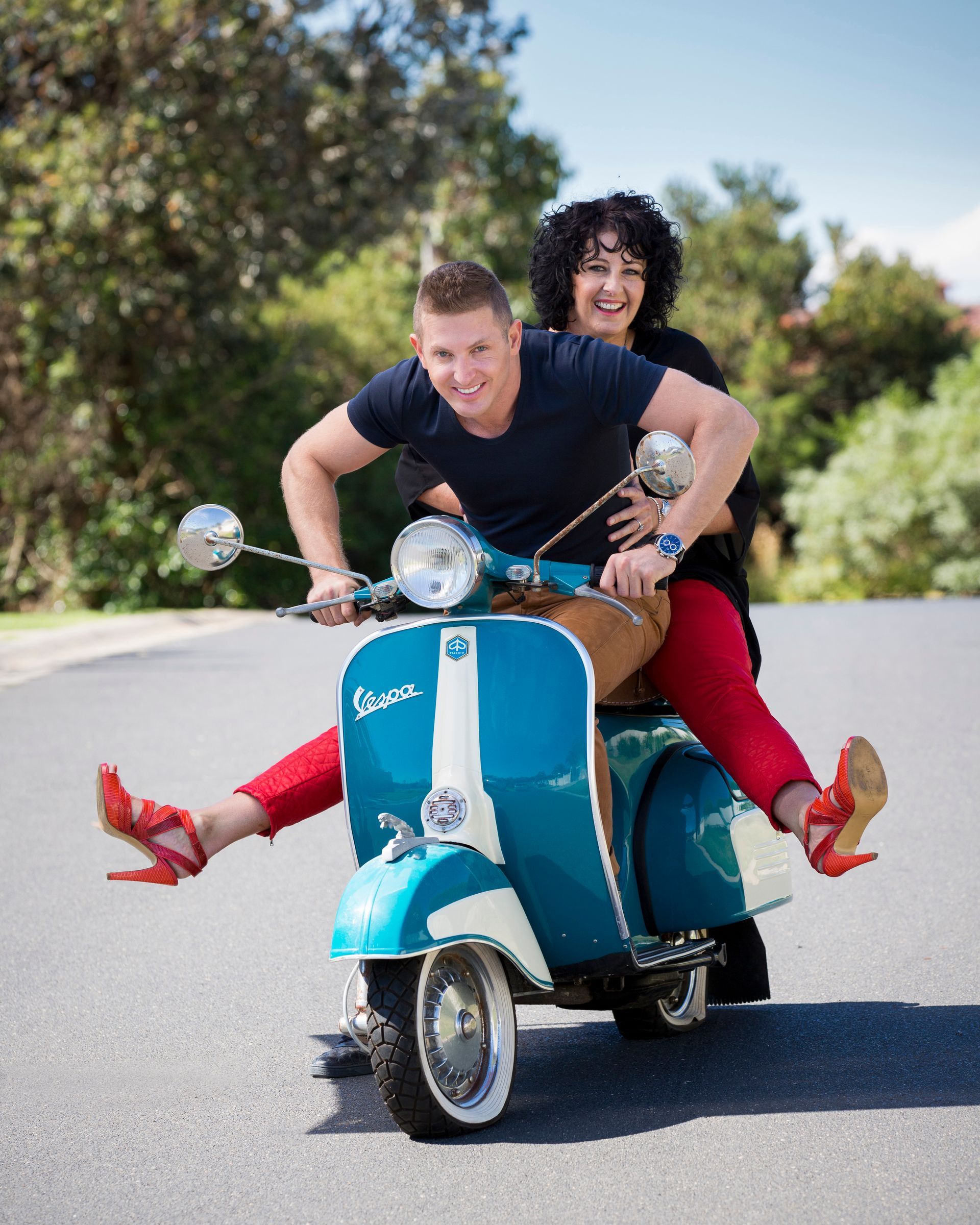 Two people riding a blue scooter down a sunny road, the passenger holding on from behind.