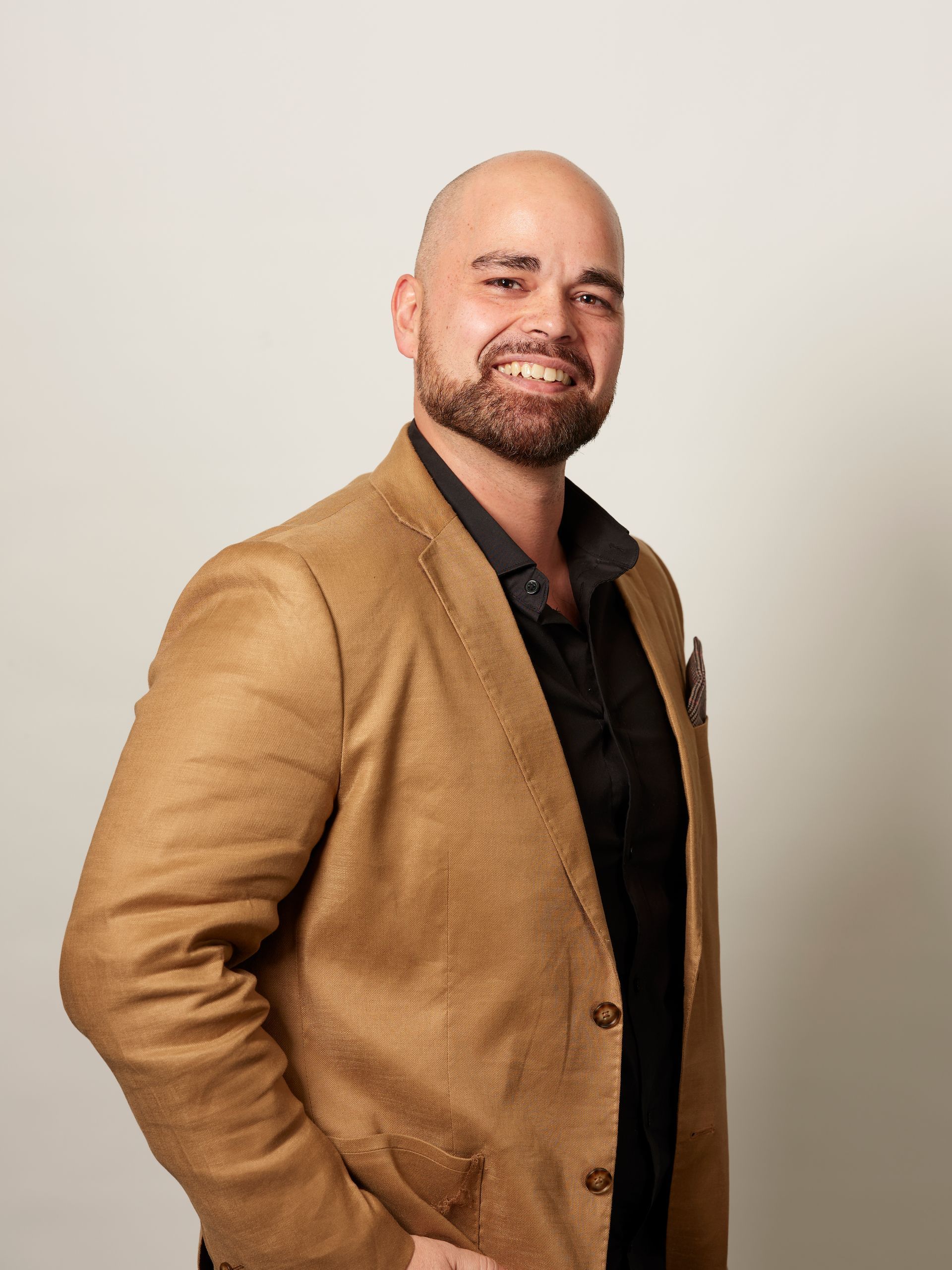 Man in a tan blazer and black shirt smiling against a plain light background
