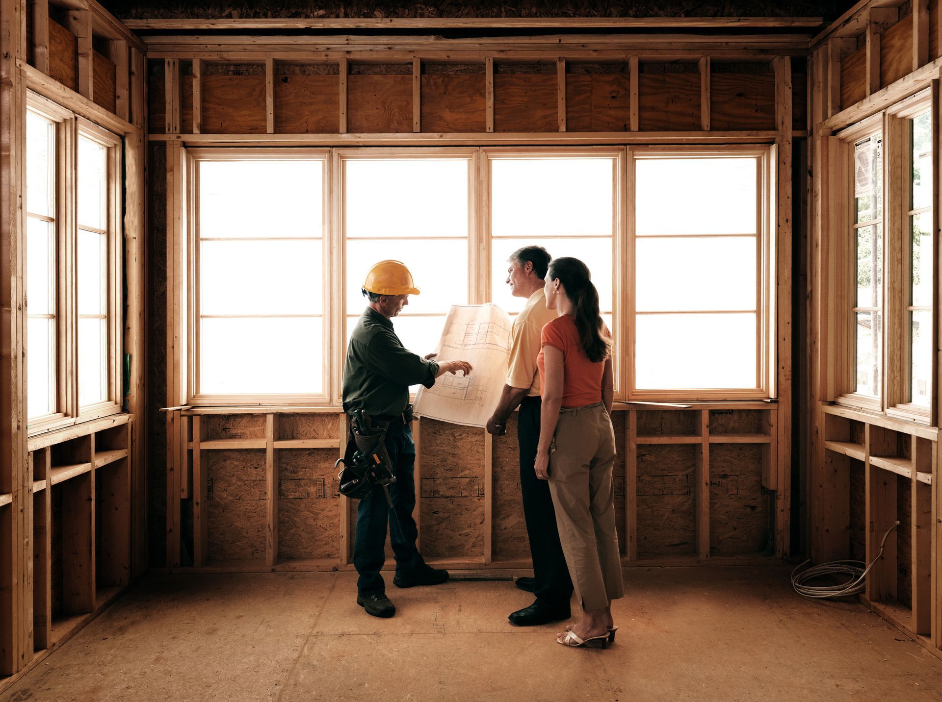 Three people in a framed house interior reviewing plans with a construction worker in a hard hat.