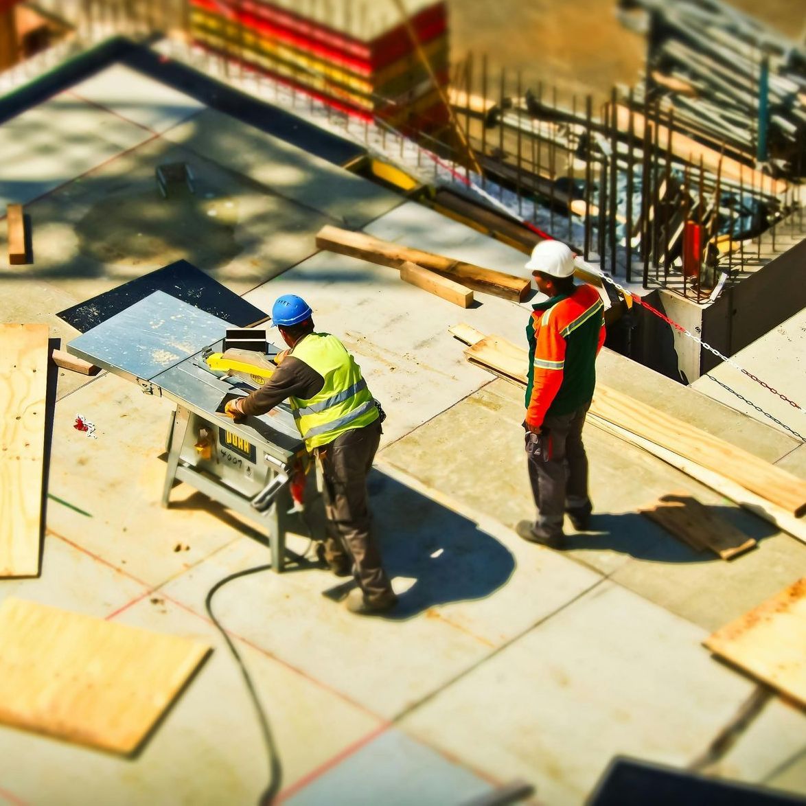 Two construction workers in safety vests and helmets cutting material on a rooftop construction site