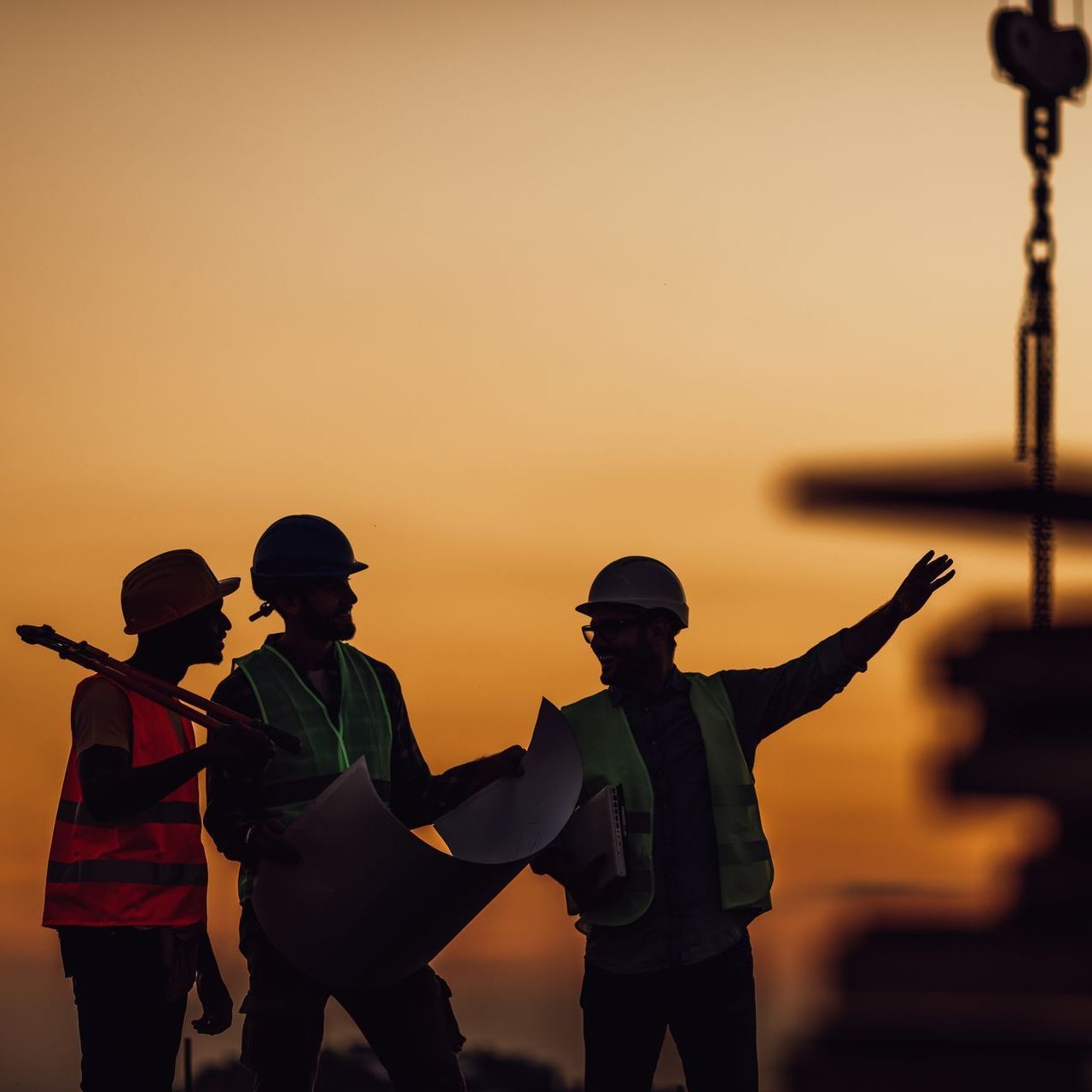Construction workers silhouetted at sunset, one pointing while holding plans near a crane