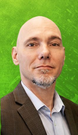 Bald man with a goatee wearing a suit jacket, in front of a green backdrop.