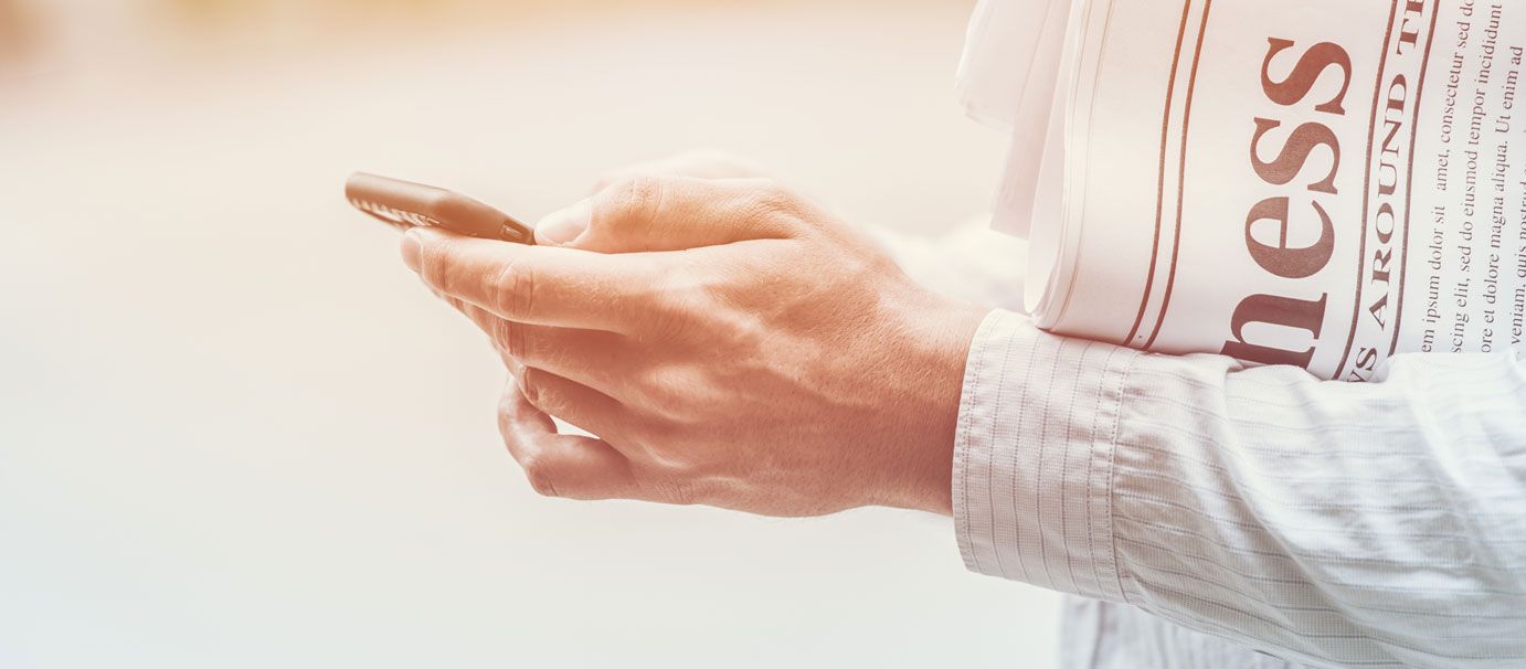 A person's hands holding a phone and a newspaper, the word 