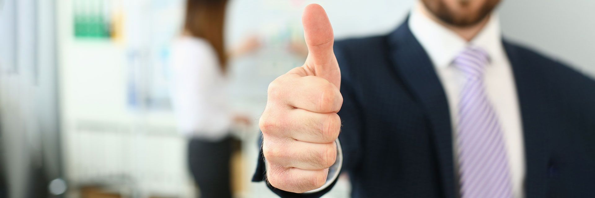 A person in a suit gives a thumbs-up. A woman in a white shirt and black pants is blurred in the background.