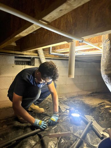 Person in a crawl space welding metal pipes under exposed beams, bright blue arc light illuminating the area.