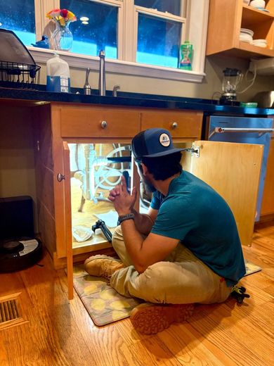 Person kneels under kitchen sink, inspecting plumbing with a flashlight in a wooden kitchen.