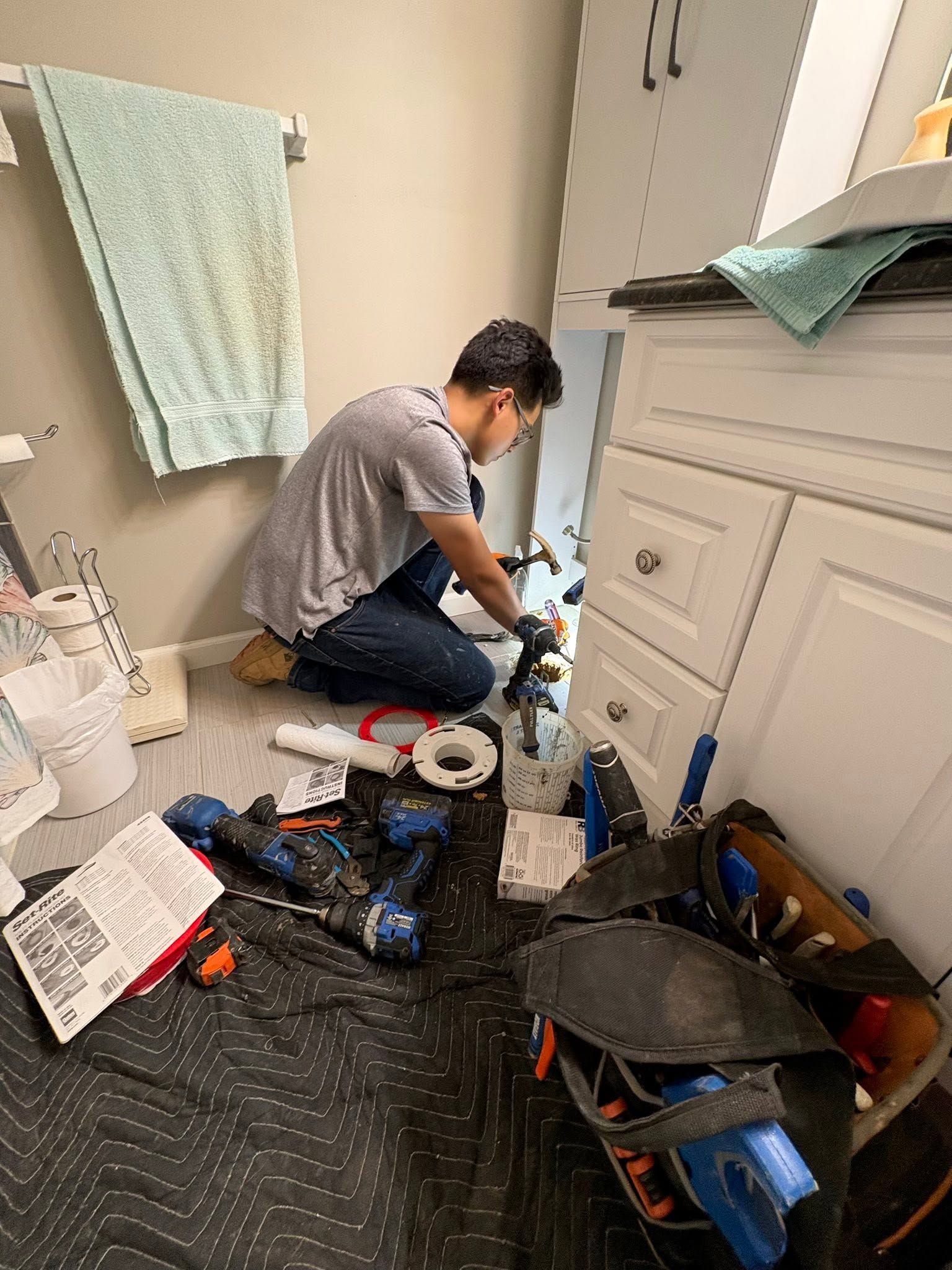 Person kneeling on a bathroom floor fixing plumbing beside a white vanity, tools scattered around.