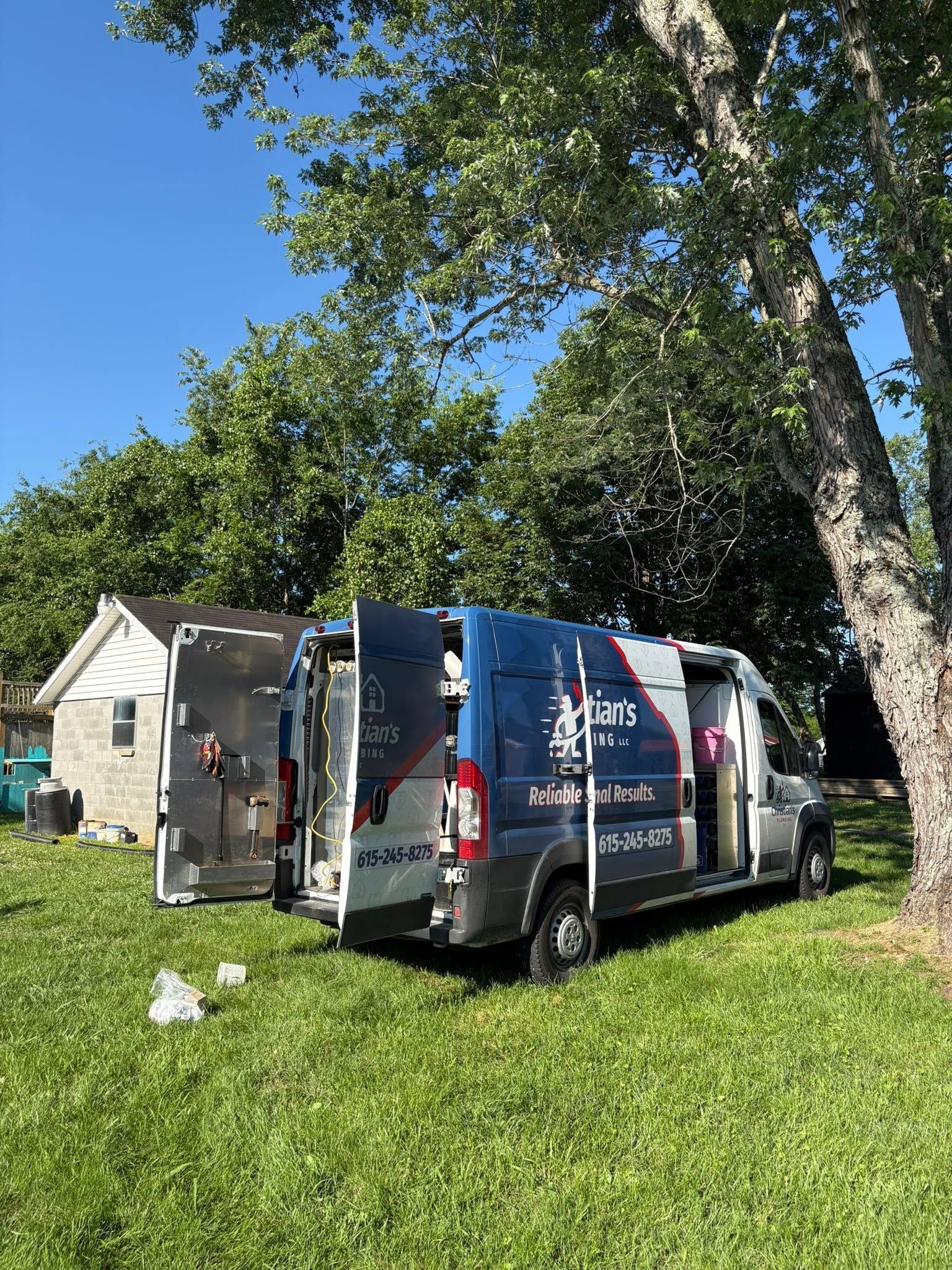 White utility van with open rear doors parked on grass beside a house under trees.