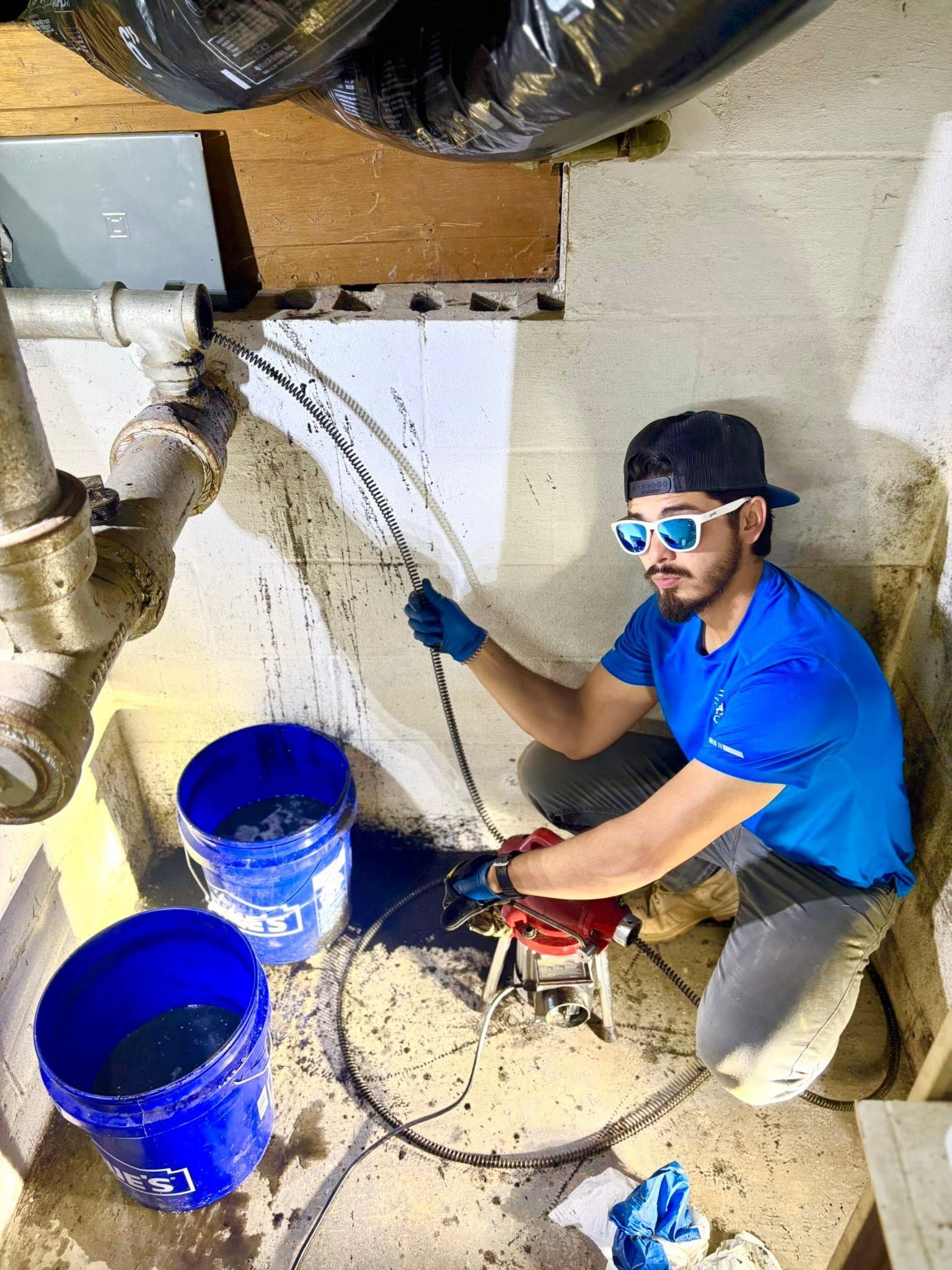 Technician in blue shirt uses a wall tool near two blue buckets during indoor repair work