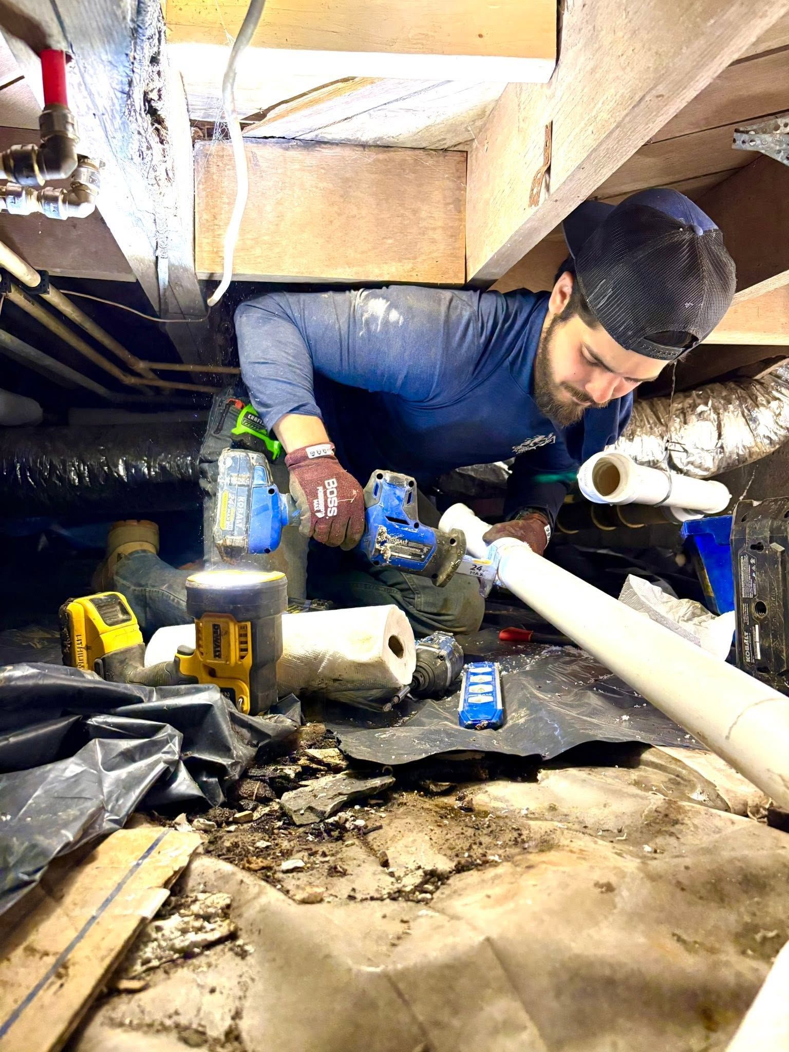 Person crawling under a house with tools and pipes in a cramped crawlspace.