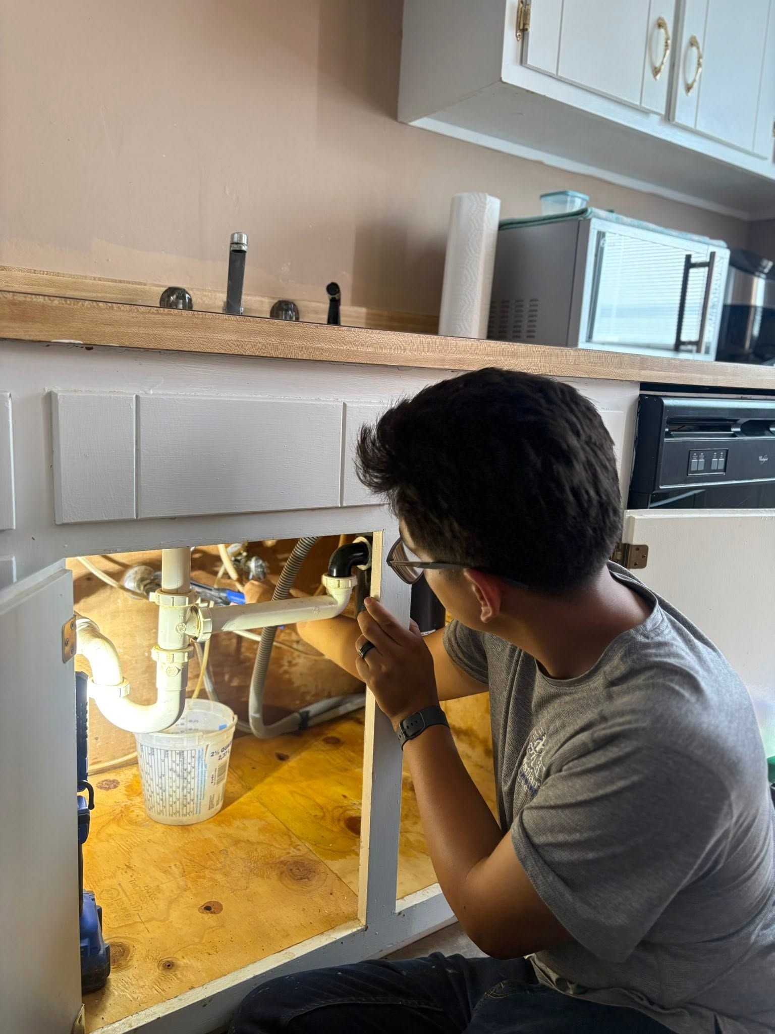 Person crouches under a kitchen sink, inspecting exposed pipes with a tool.