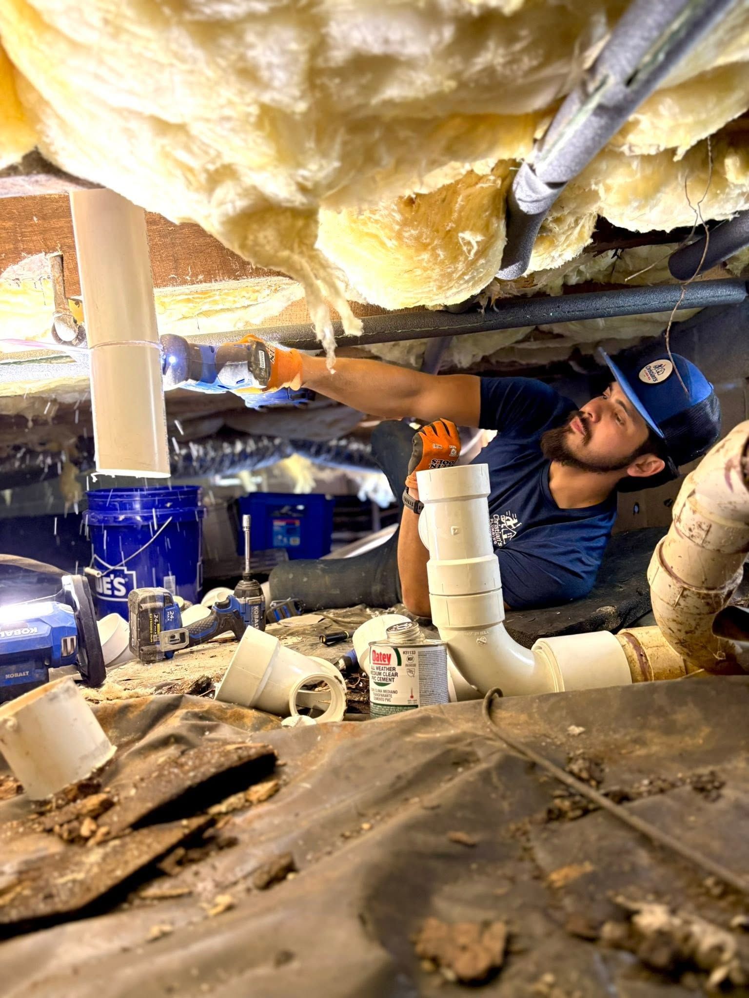 Plumber under a sink repairing PVC pipes with tools, surrounded by insulation and debris