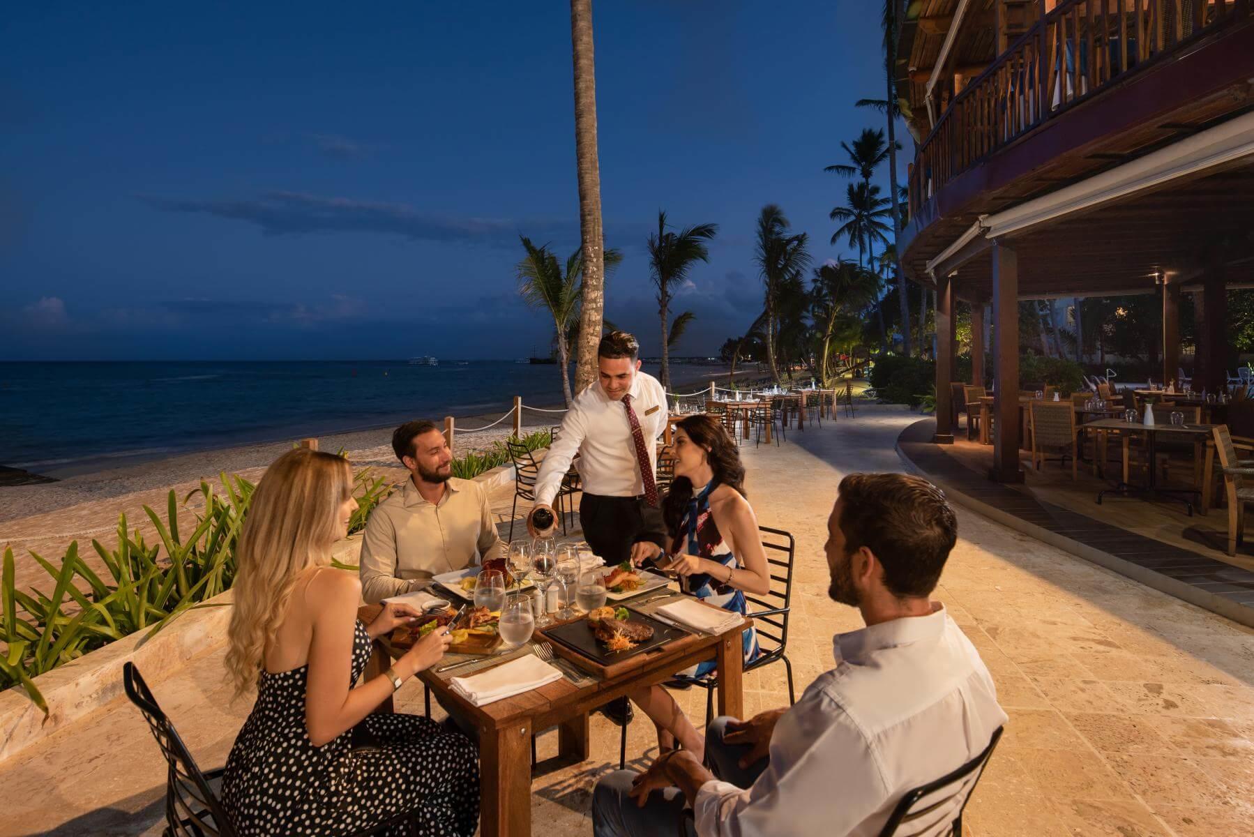 Un grupo de personas está sentada en una mesa en la playa comiendo.