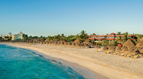 Una playa con sombrillas de paja y un hotel al fondo.