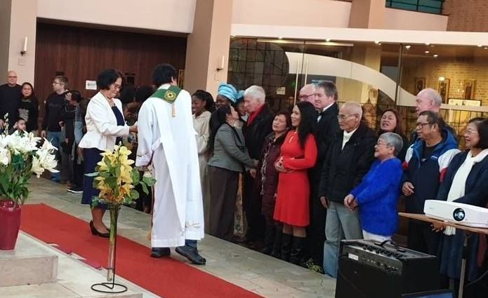 People greeting a robed person on a red carpet in a church. Guests watch, some smiling. Flowers, windows visible.