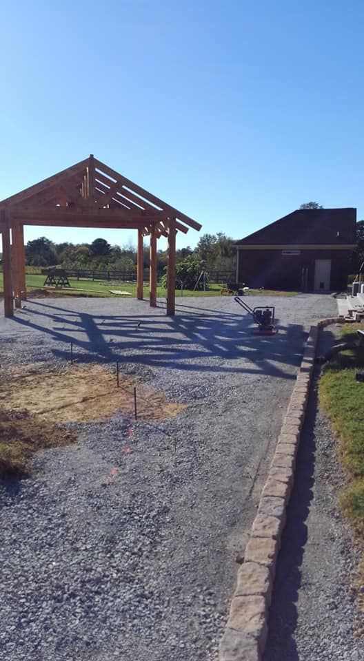 A wooden pavilion is sitting in the middle of a gravel driveway next to a house.