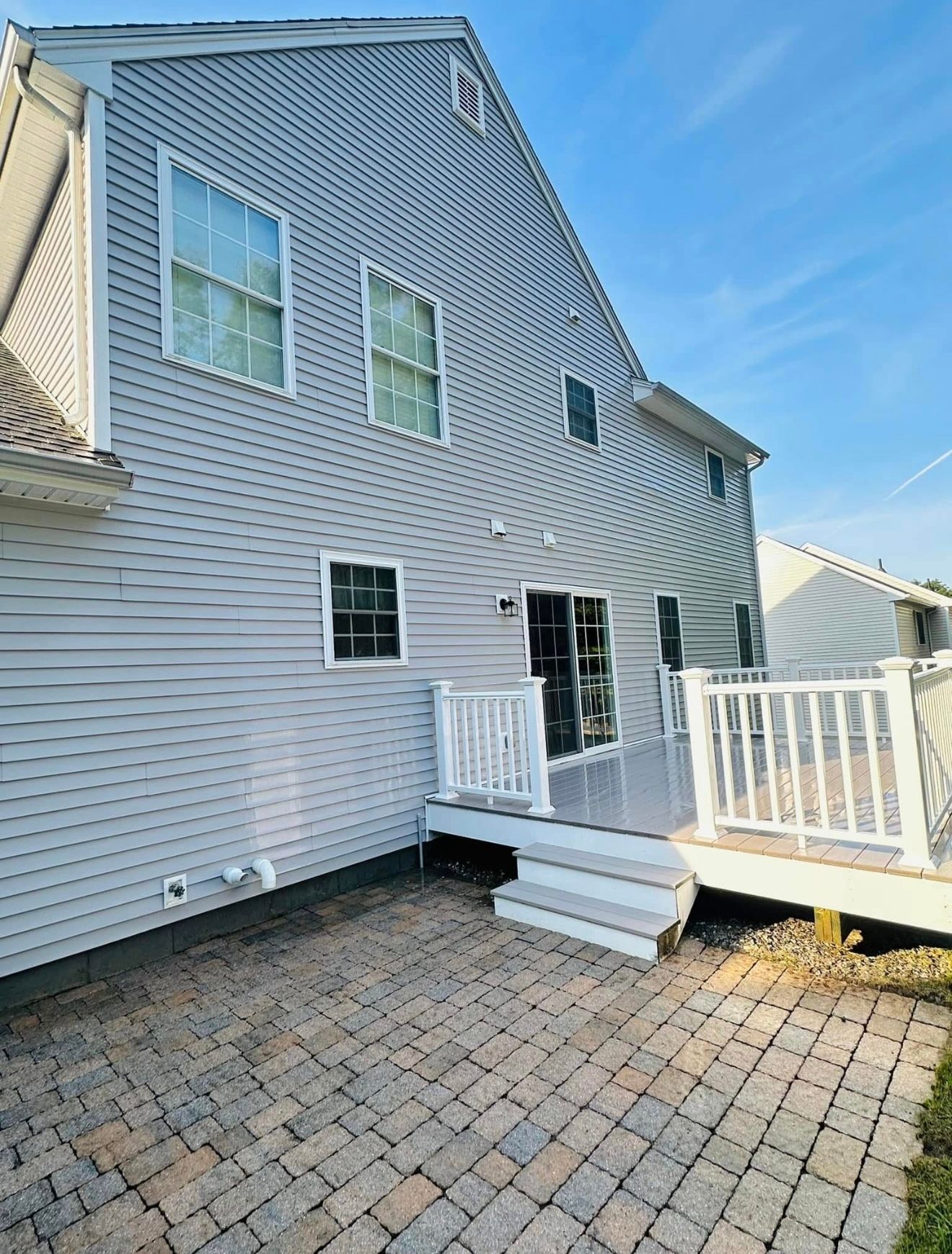 Back of a two-story house with gray siding, deck, and patio. Blue sky.