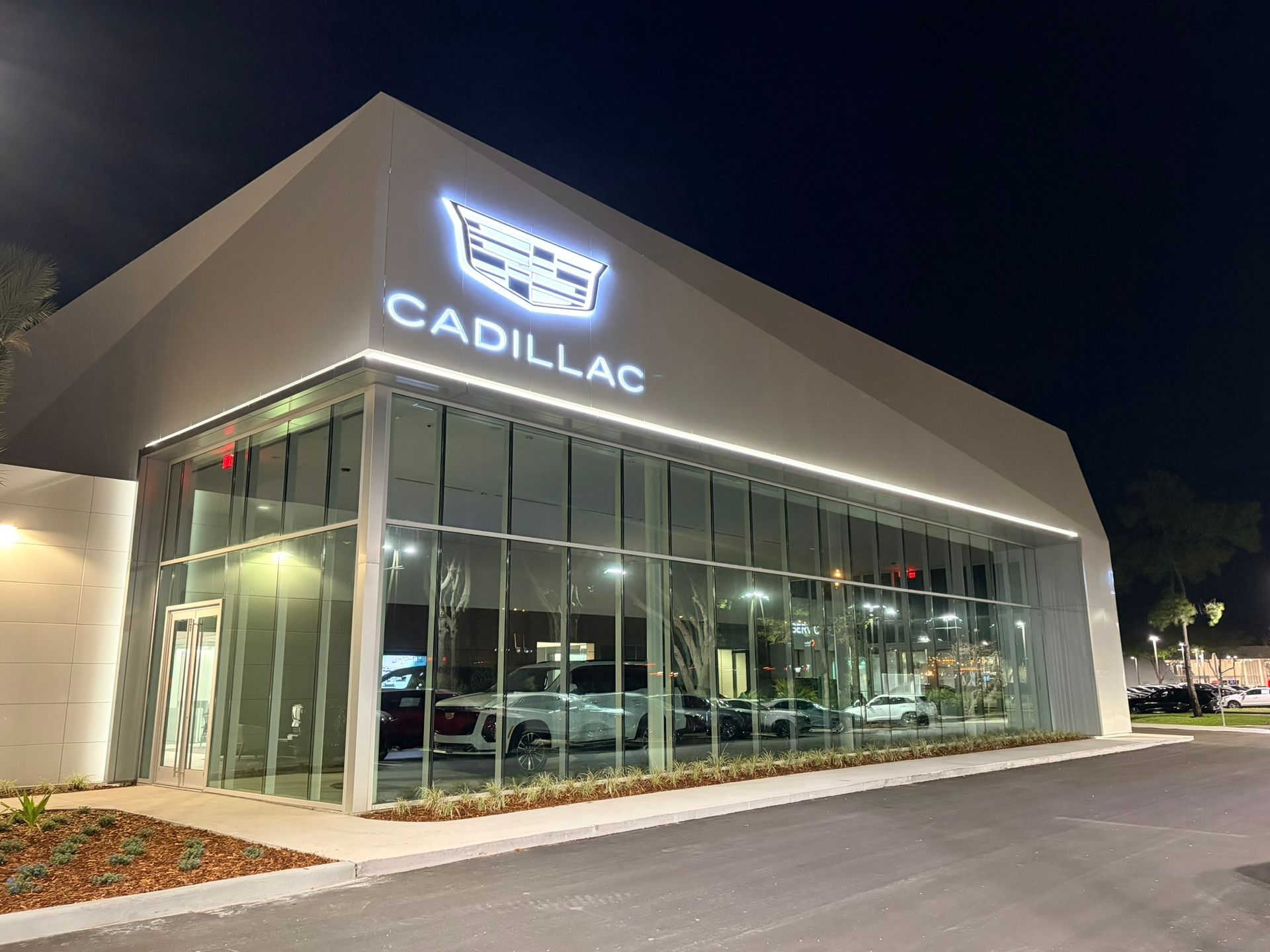 Modern Cadillac dealership showroom at night, illuminated by interior lights and a glowing white brand sign on the exterior.