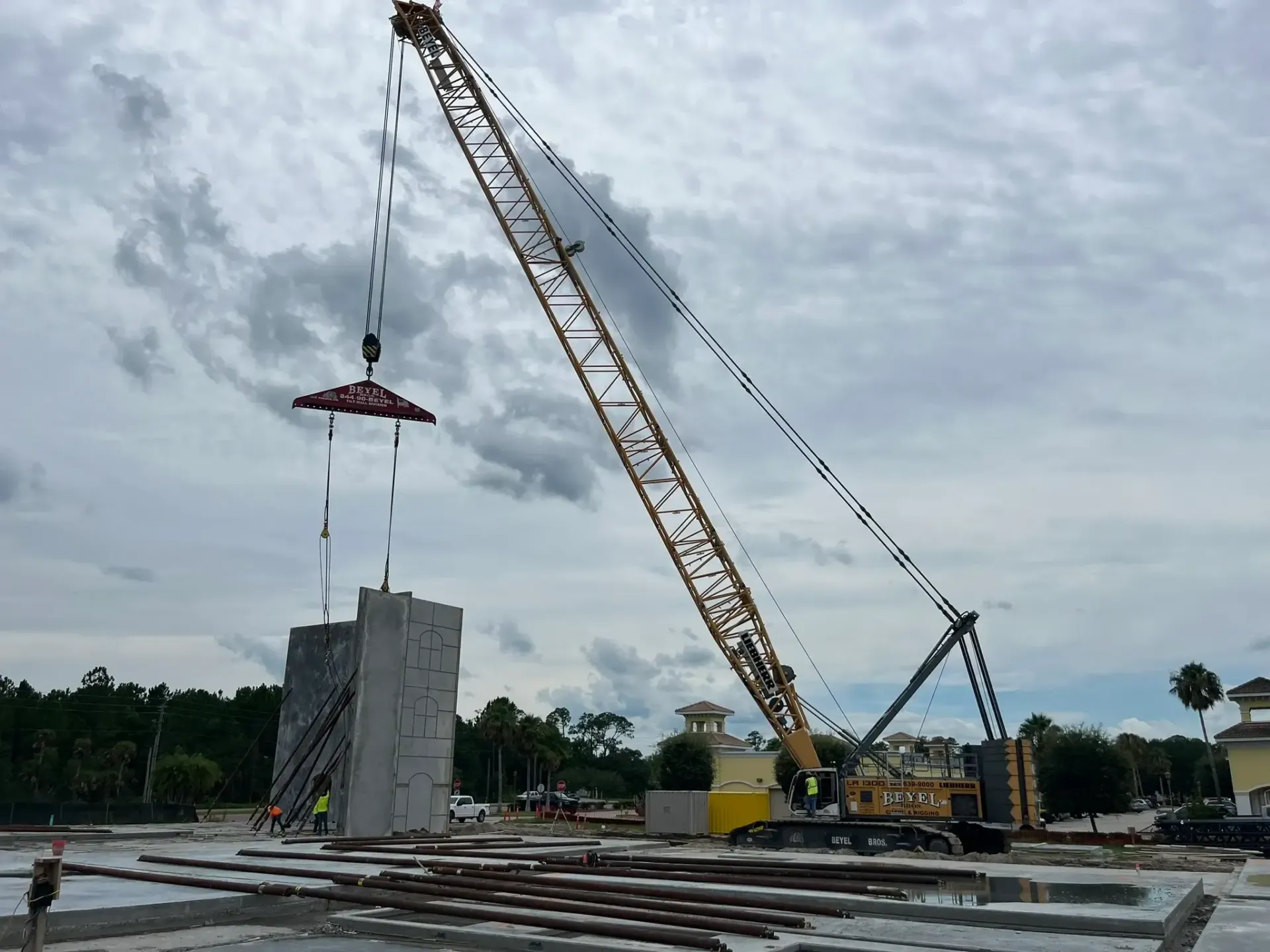 Crane lifting a large tilt-wall concrete panel near a building under construction in Florida, cloudy sky above.