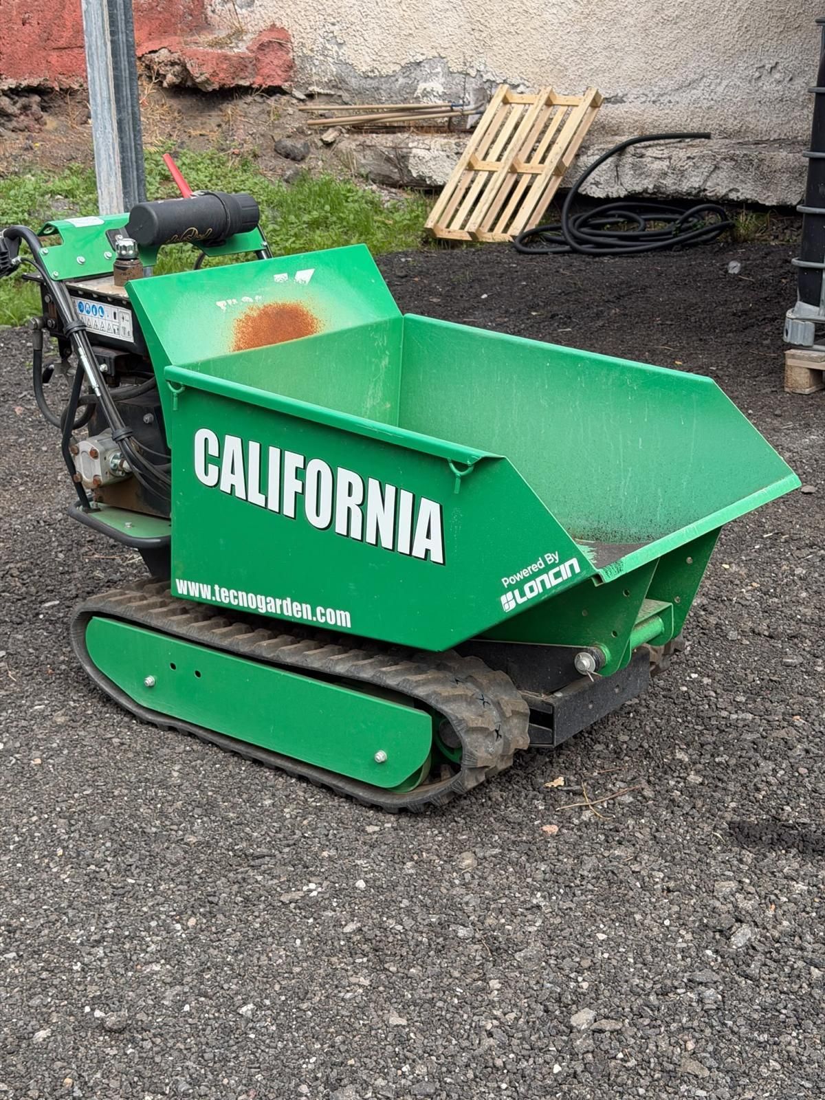 Mini dumper cingolato verde con marchio California, su ghiaia accanto a un muro e a pallet di legno