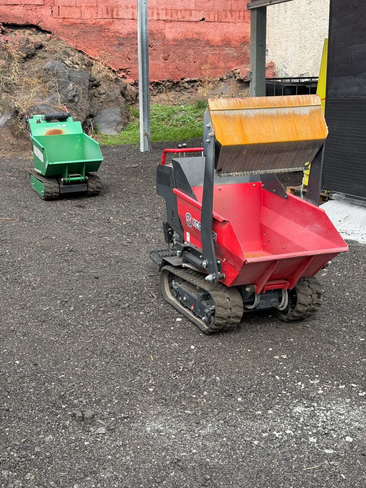 Due piccole macchine da cantiere cingolate, una rossa e una verde, parcheggiate sulla ghiaia vicino a un muro.