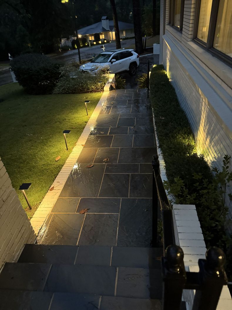 Rainy nighttime walkway beside a lit house, with a white car parked ahead and wet stone steps in the foreground.