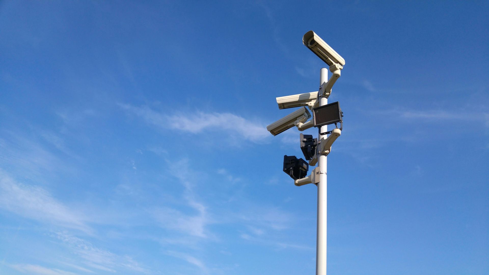Surveillance cameras and floodlights atop a tall white pole against a blue sky.