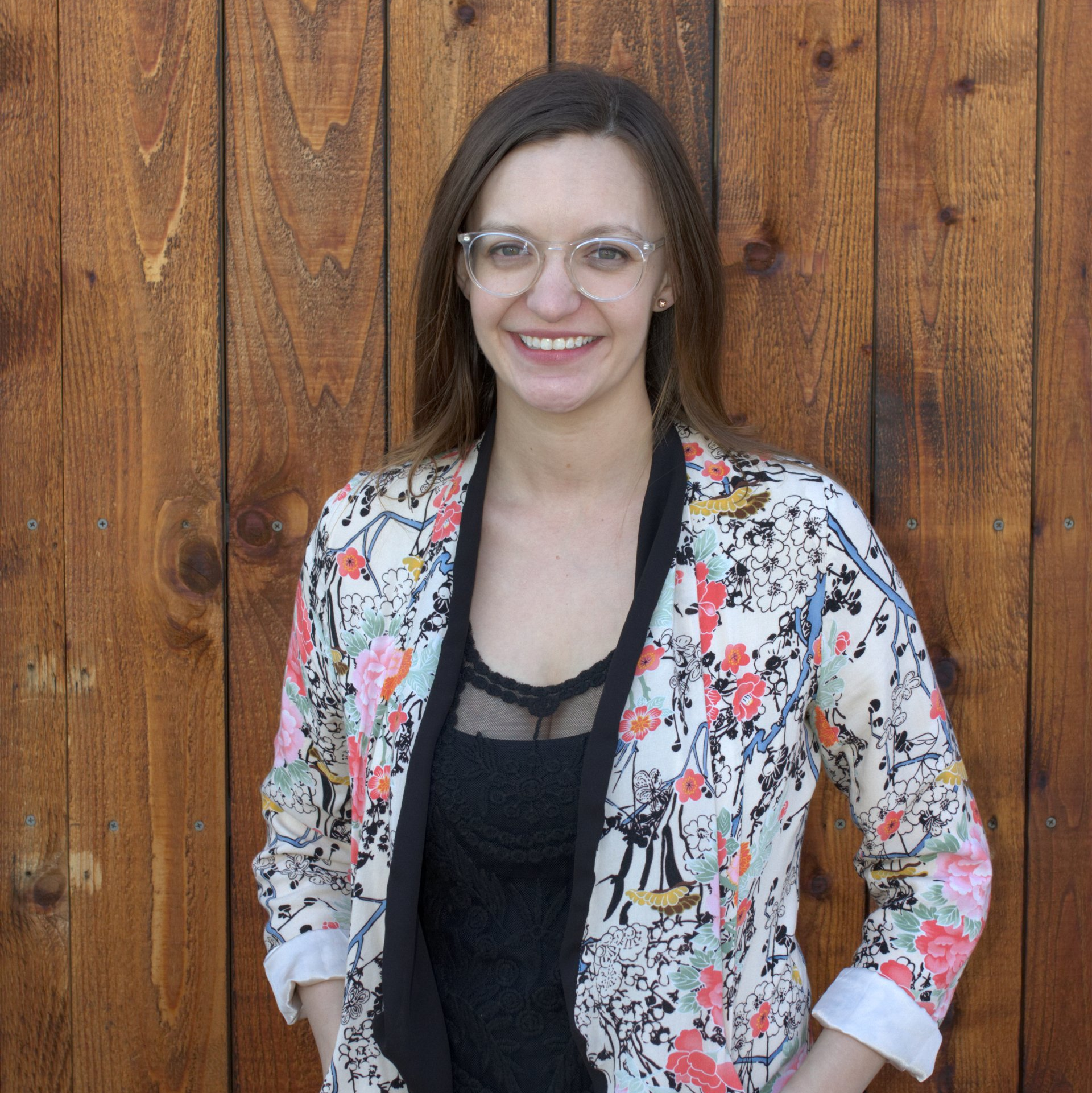 Woman in glasses, patterned jacket, smiling in front of a wood wall.