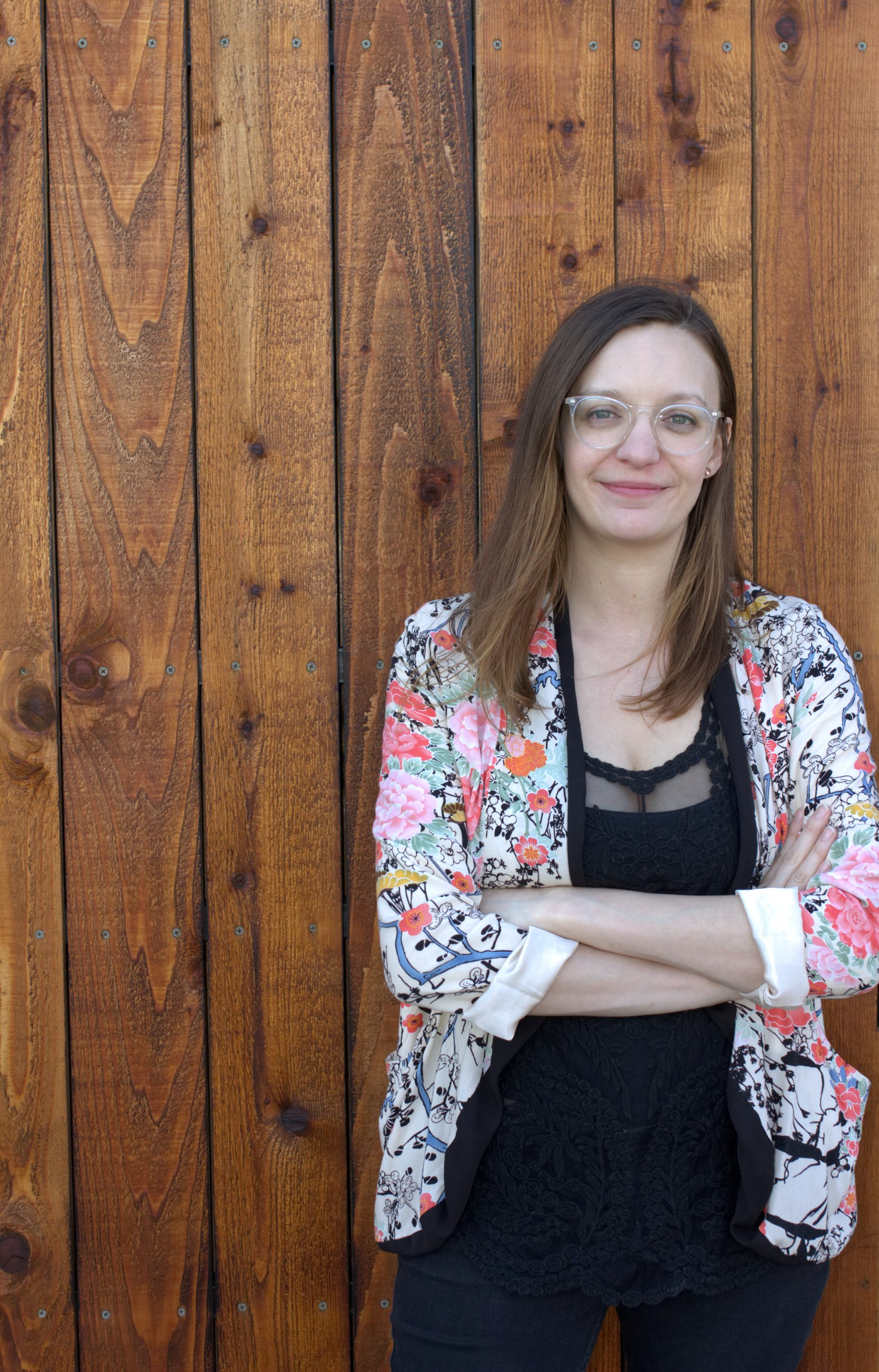 Woman with glasses and floral jacket, arms crossed, stands against a brown wood wall.