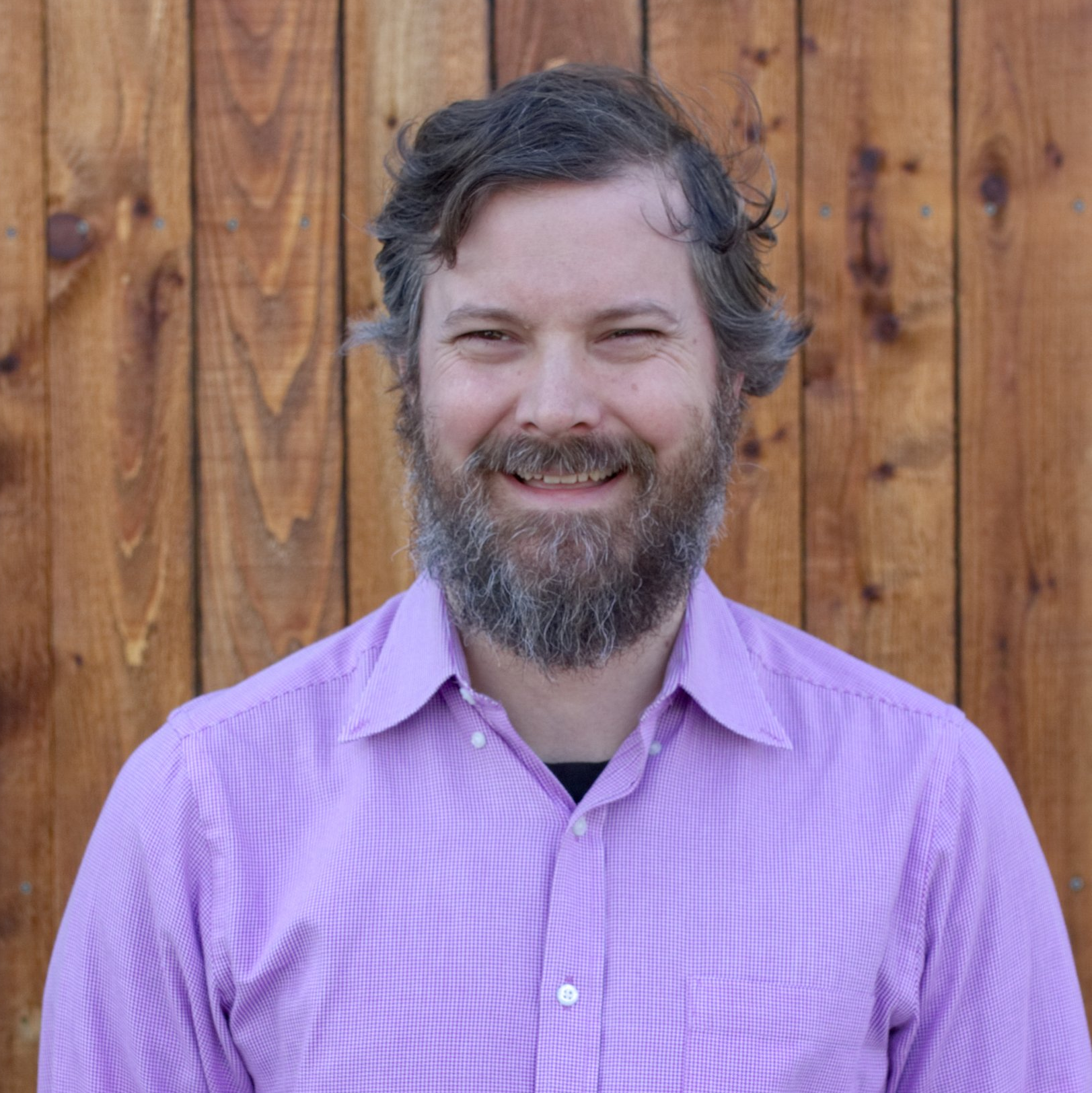 Man with a beard smiling in a pink collared shirt against a wooden wall.