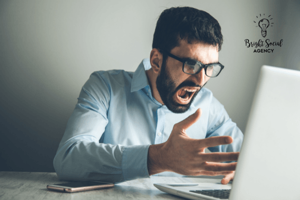 Man in glasses, yelling, and gesturing at a laptop; looks frustrated, indoors.