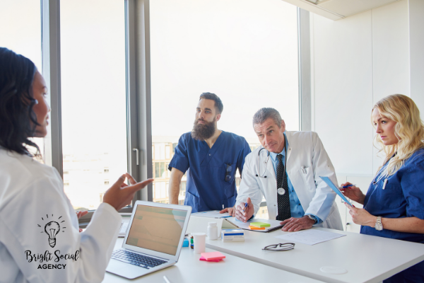 Medical professionals in a meeting; woman points, others look at documents, near a window.