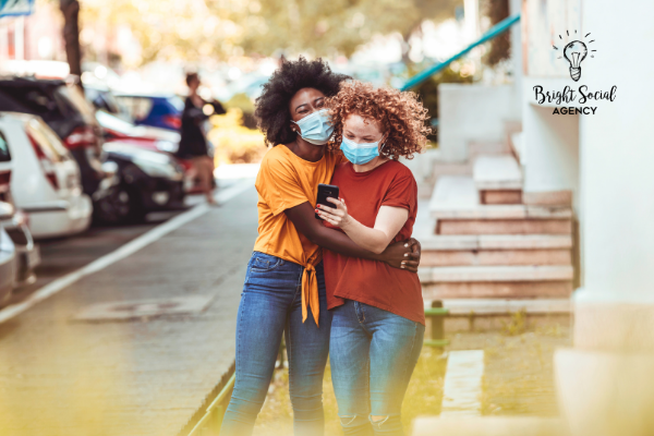 Two people wearing masks hugging, looking at a phone on a city street.